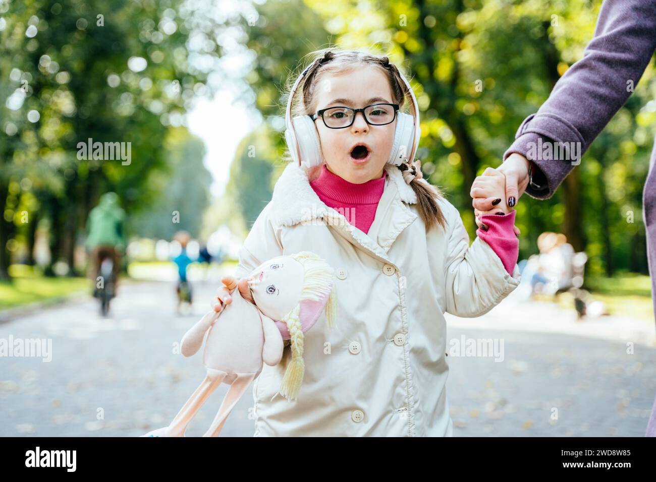 Cute little girl with special needs singing, listening music with headphones holding toy and her mother's hand in the outdoor park. Stock Photo