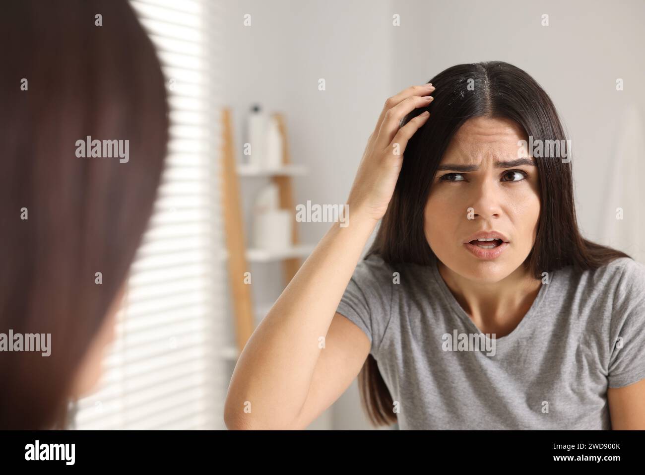 Emotional woman examining her hair and scalp near mirror in bathroom. Dandruff problem Stock Photo