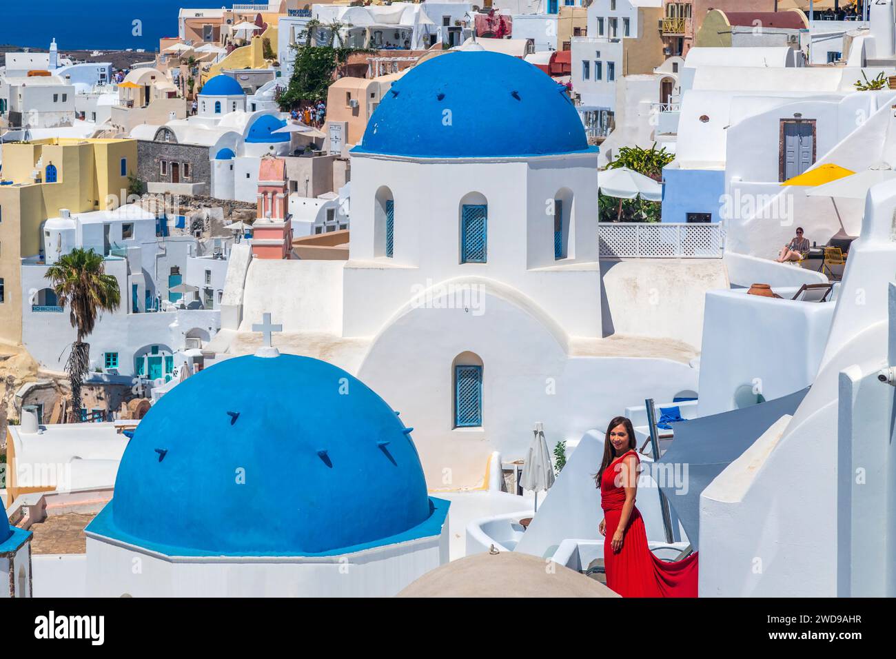 Oia, Santorini island, Greece - June 21, 2021: Beautiful girl in red dress and typical white-blue traditional architecture with domes and churches. Stock Photo