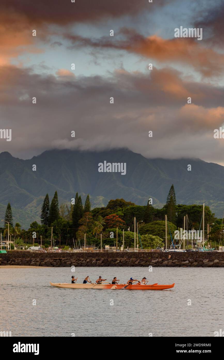 A traditional multi-person outrigger boat being paddled in the sea in Hawaii. Behind the ocean is a mountain side, with low clouds Stock Photo