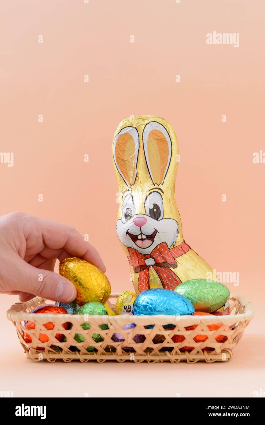 Detail of a hand picking up a chocolate Easter egg from a basket full of chocolate eggs, next to a chocolate bunny with a background of a soft orange Stock Photo