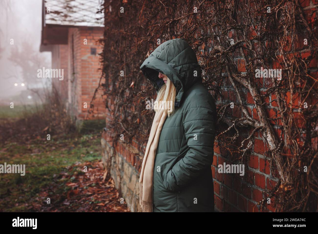 Sad lonely woman with hood standing by brick wall in fog. Loneliness and depression in own thoughts Stock Photo
