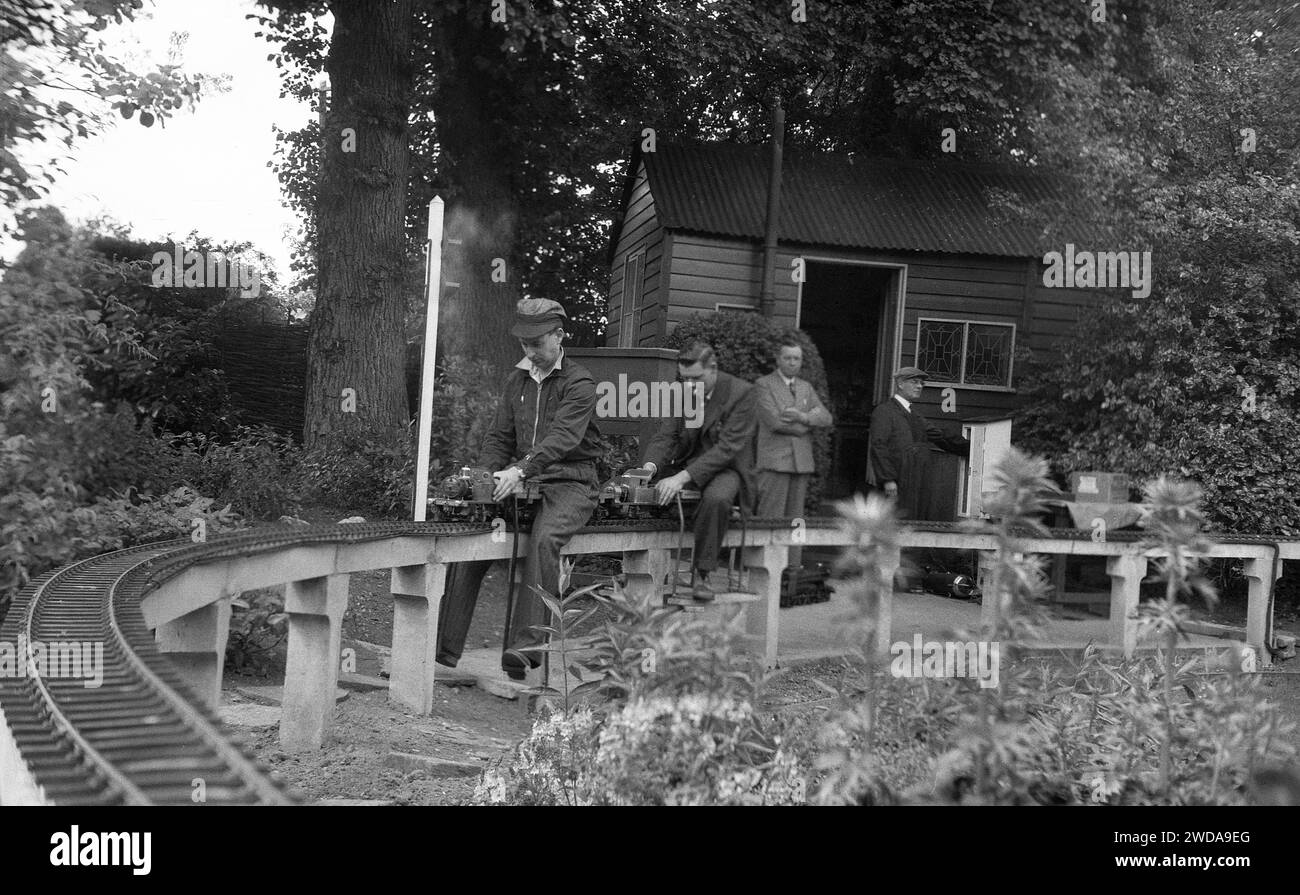 1950s, historical, outside in a garden, model railway enthusiasts at a miniature garden steam railway, at the Field End Railway Co, England, UK.  On a raised track sitting on concrete posts, two men riding on the tiny steam trains, one being a scaled model of the GWR 'County of Oxford' (1023 ) steam locomotive, the original of which started service in 1947. Stock Photo
