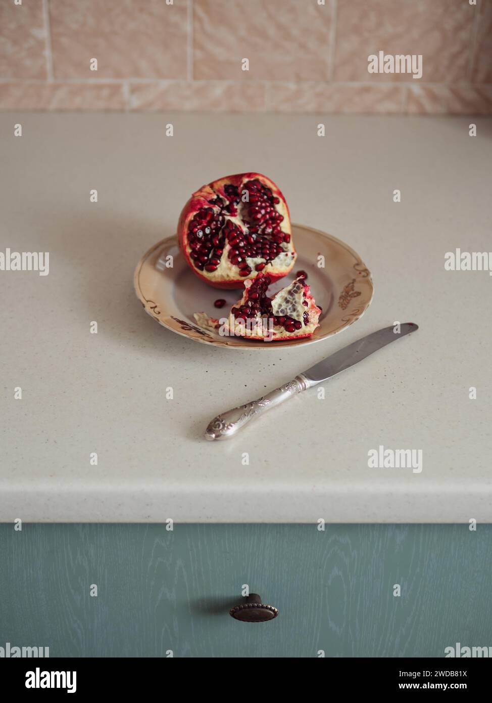 Vertical close up view of an open pomegranate on a plate against the white table and pastel pink wall. A part of turquoise coloured drawer in the fram Stock Photo