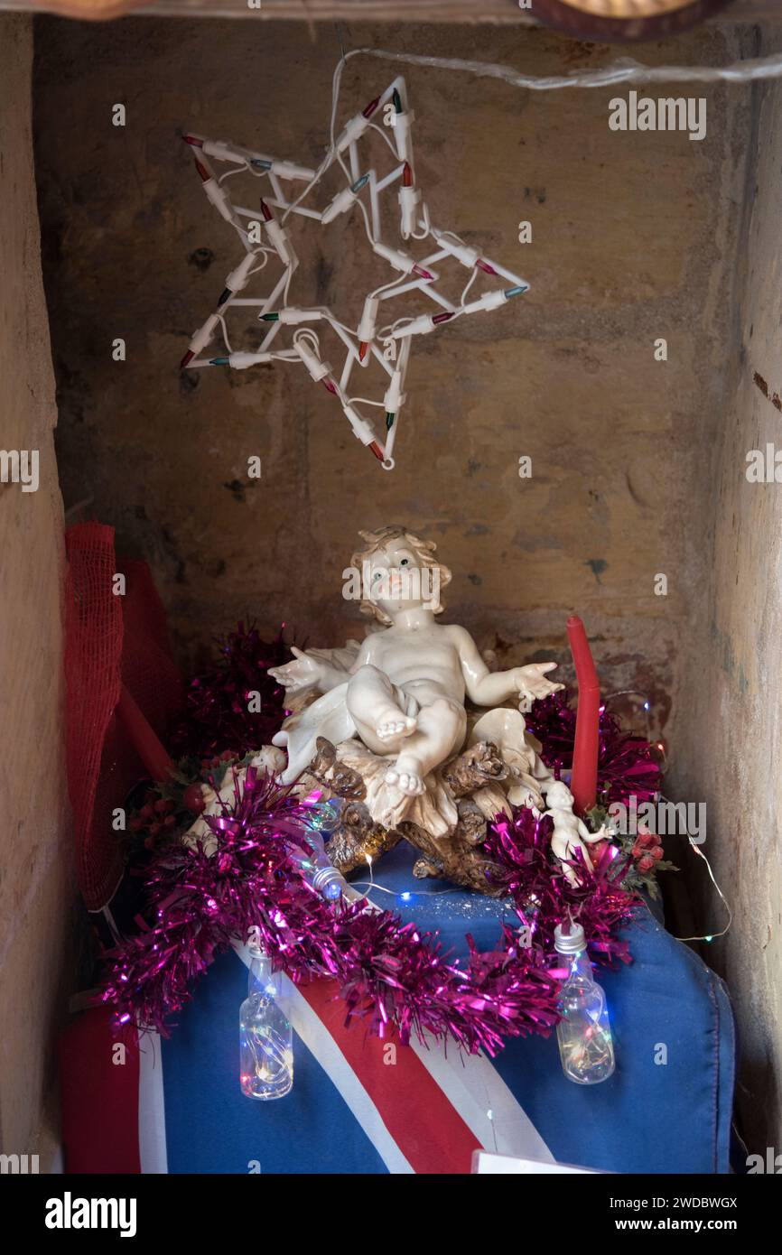 Catholic art an inexpensive ceramic ikon of the Christ Child lying on a Union Jack flag in a nook in an old shop. A shrine in the family store.  The lights around it flash on and off. Valletta, Malta, Island of Malta. 2024, 2020s  HOMER SYKES Stock Photo