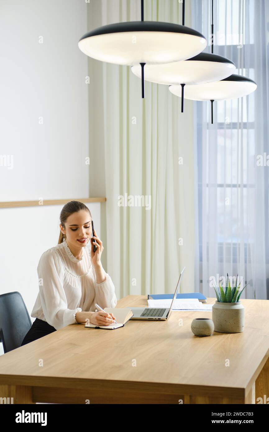 Young woman in office making notes while talking on smartphone Stock Photo