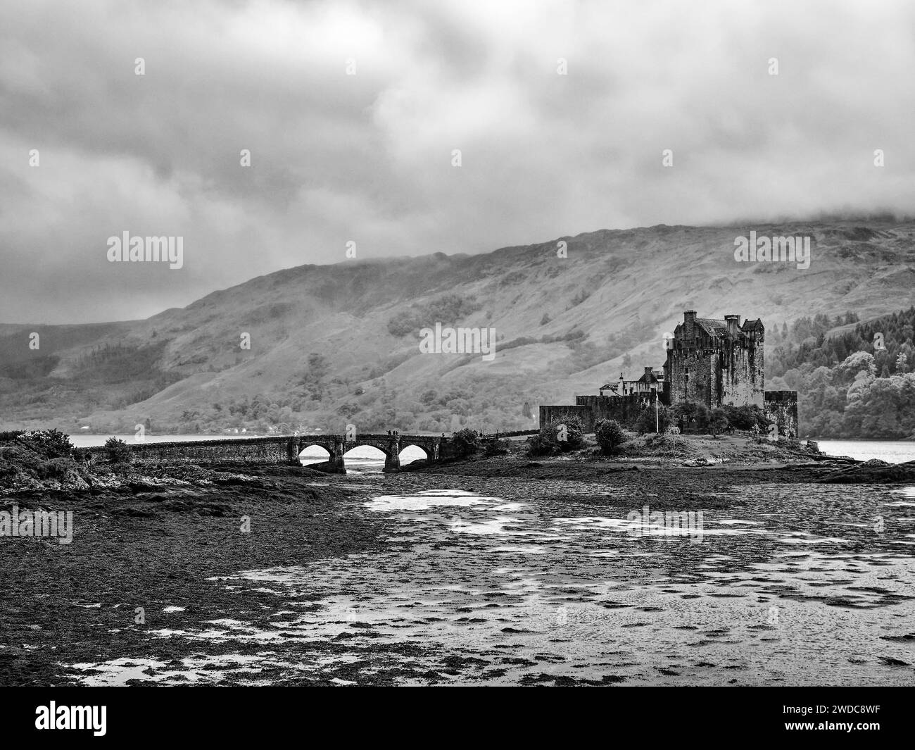 Black and white photo of an old castle with bridge in front of a mountainous landscape and cloudy sky, Eilean Donan Castle. Scotland, Great Britain Stock Photo