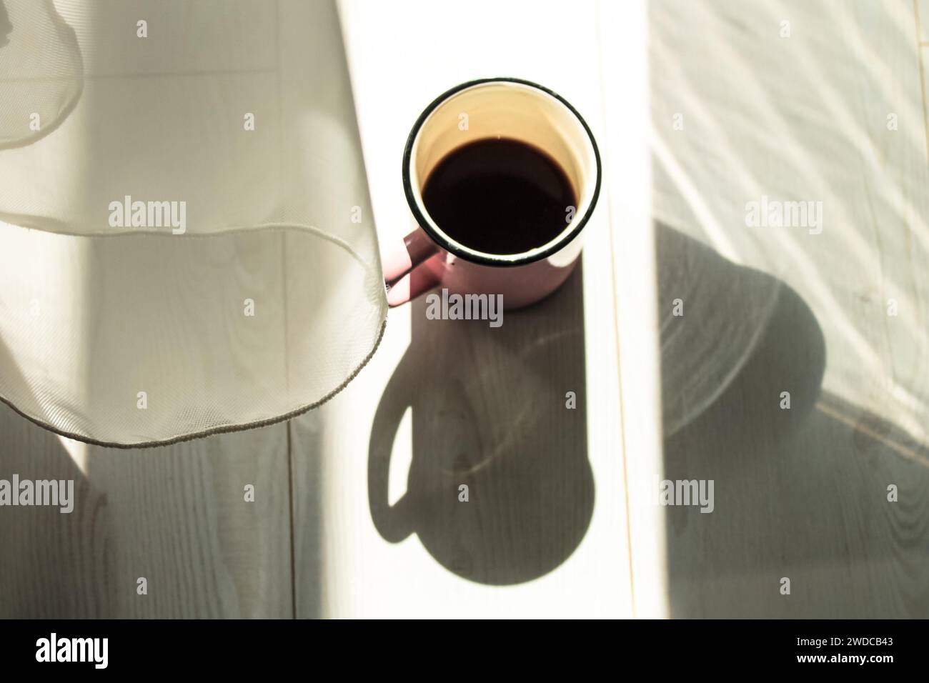 A pink cup, mug of black coffee on a white wooden parquet, contrasting long shadows on a floor in the morning light. A hot beverage for drink. Good mo Stock Photo