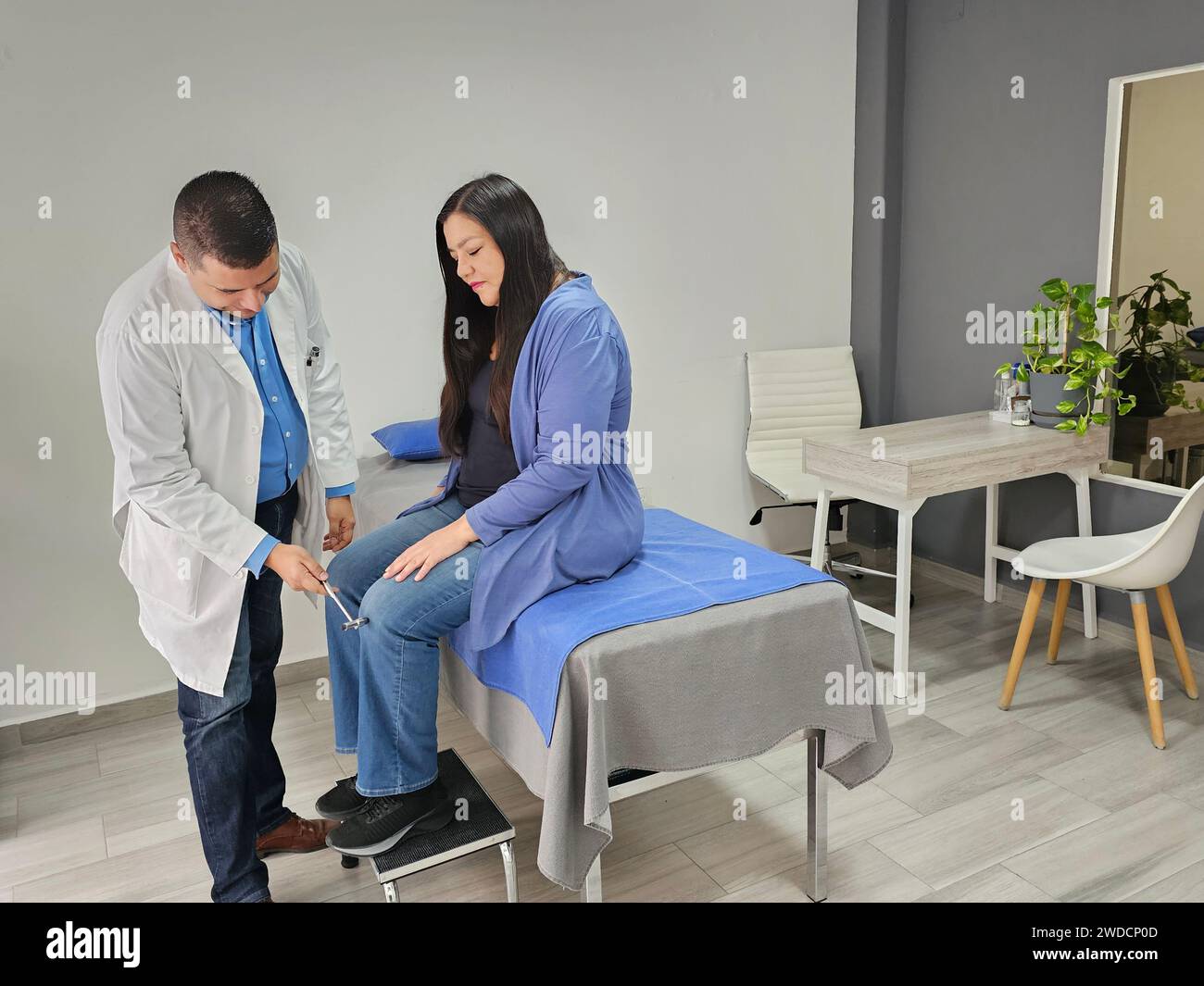 Dark-skinned Latino male doctor attends to female patient in his office initial checkup of deep reflexes with hammer Stock Photo
