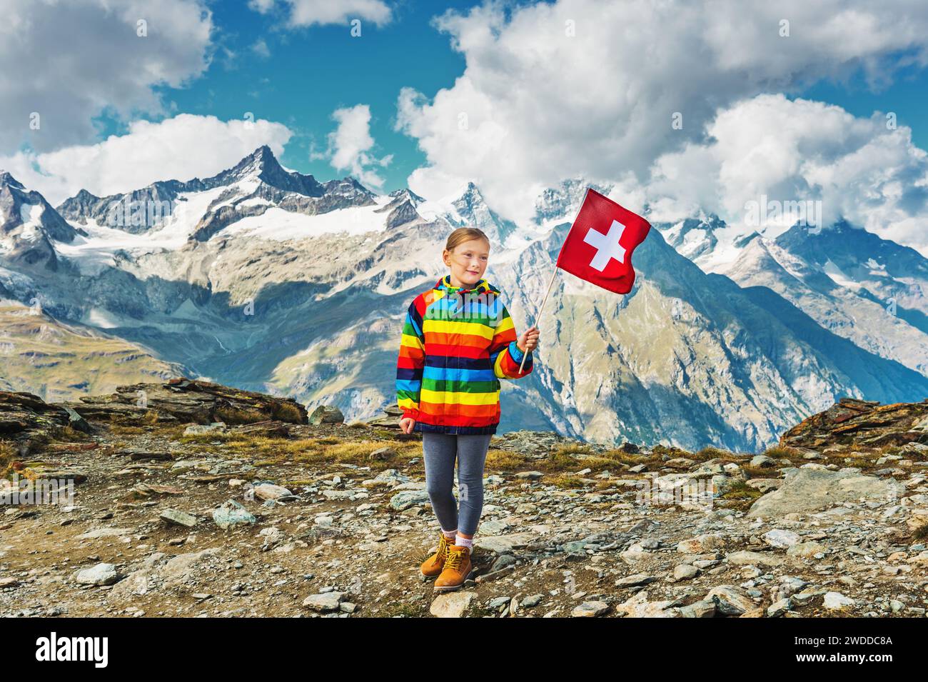 Cute little girl wearing bright rainbow colored coat, holding swiss flag, standing in front of Gornergrat glacier, Switzerland Stock Photo