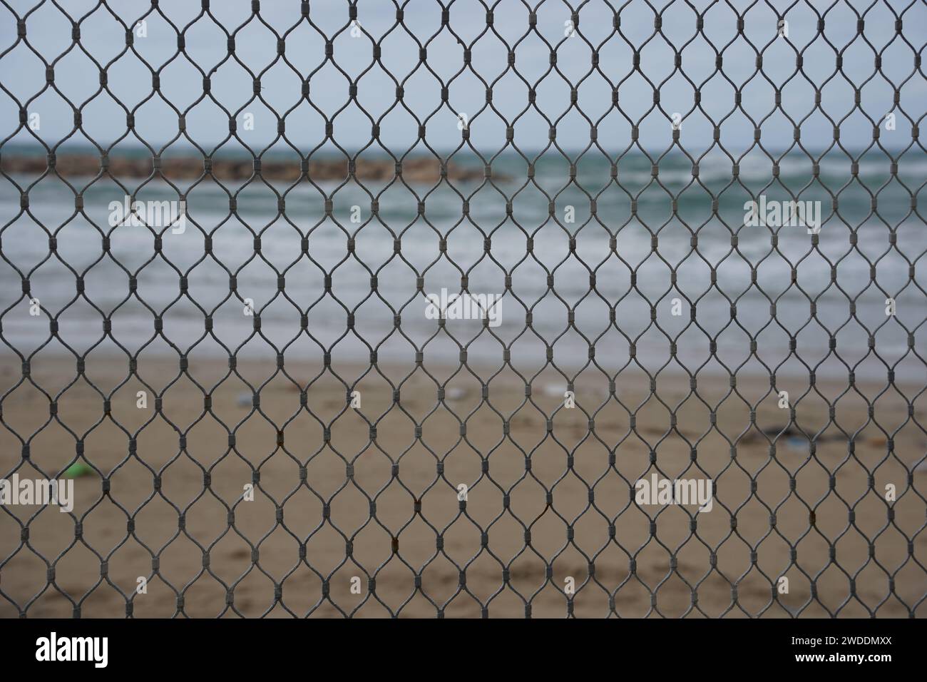 View of the winter beach through a mesh net fence. Mediterranean coast from behind metal wire fence. Stock Photo