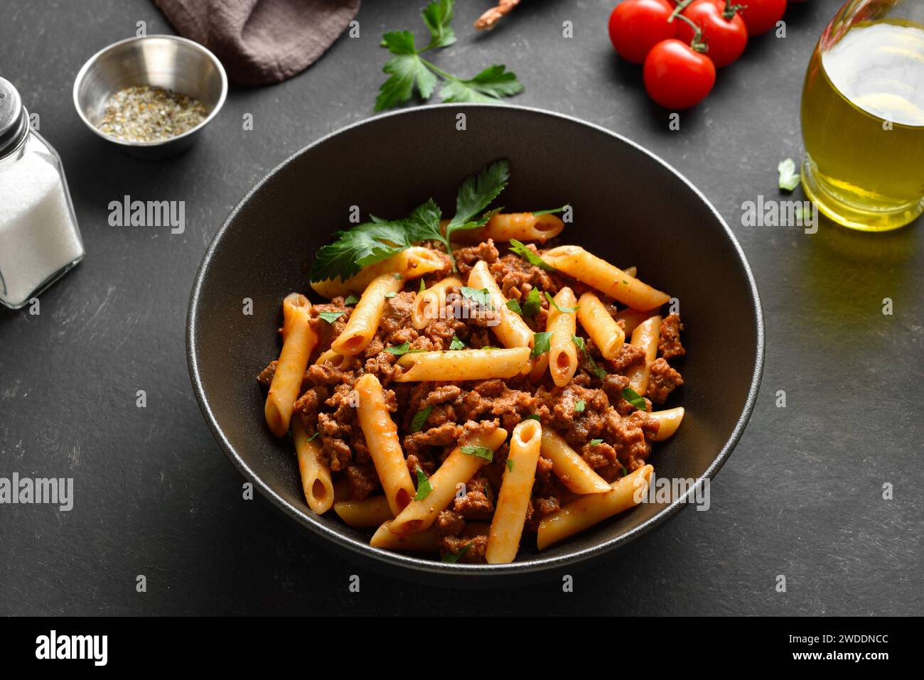 Penne pasta with minced meat, tomato sauce and greens in black ceramic bowl over dark stone background. Bowl of pasta bolognese. Close up view Stock Photo