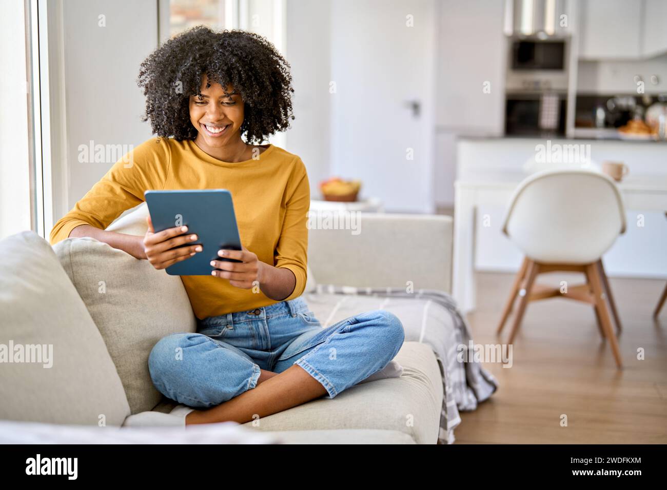 Happy young African American woman sitting on sofa at home using digital tablet. Stock Photo