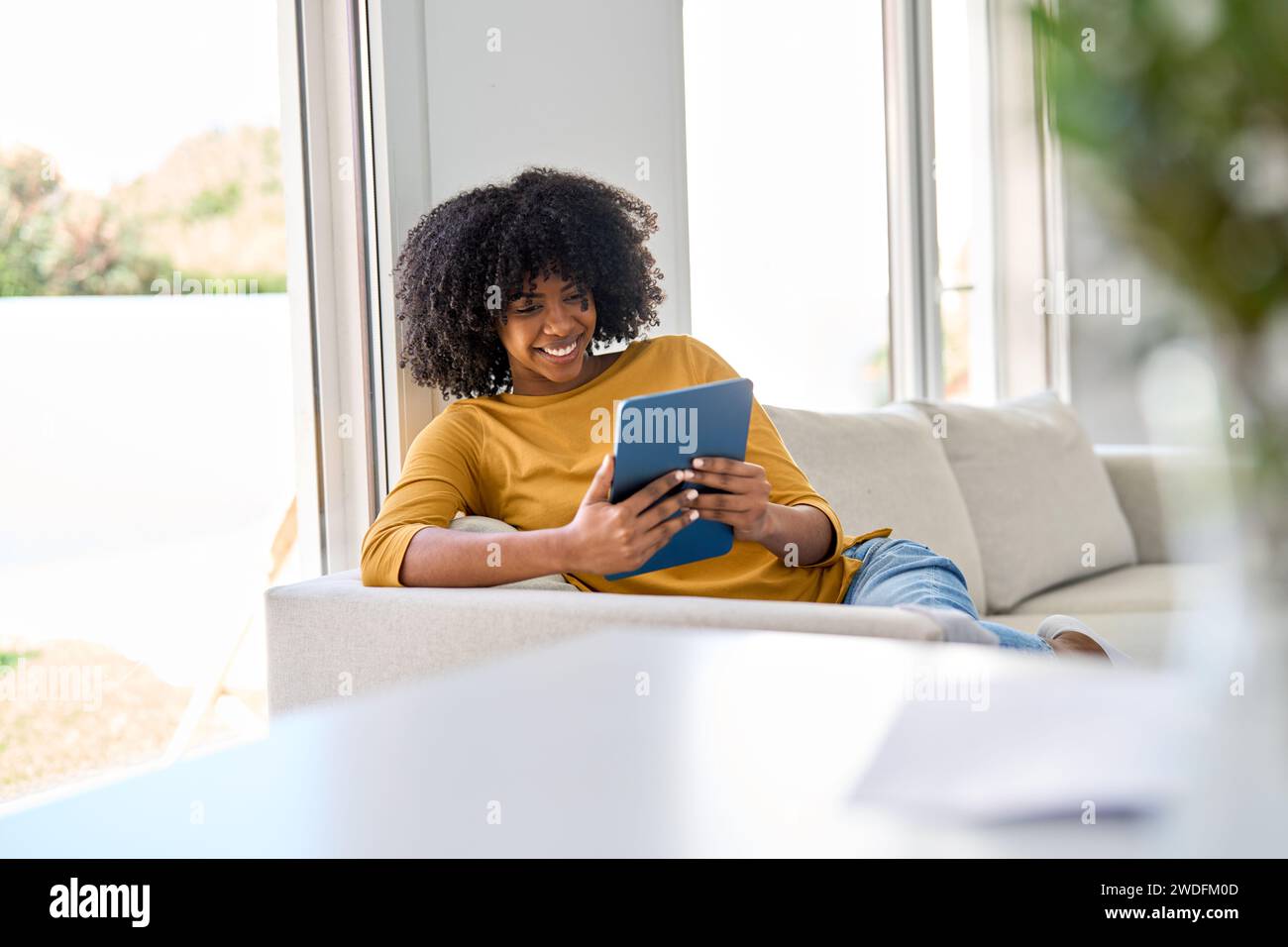 Smiling young African woman using digital tablet relaxing on couch at home. Stock Photo