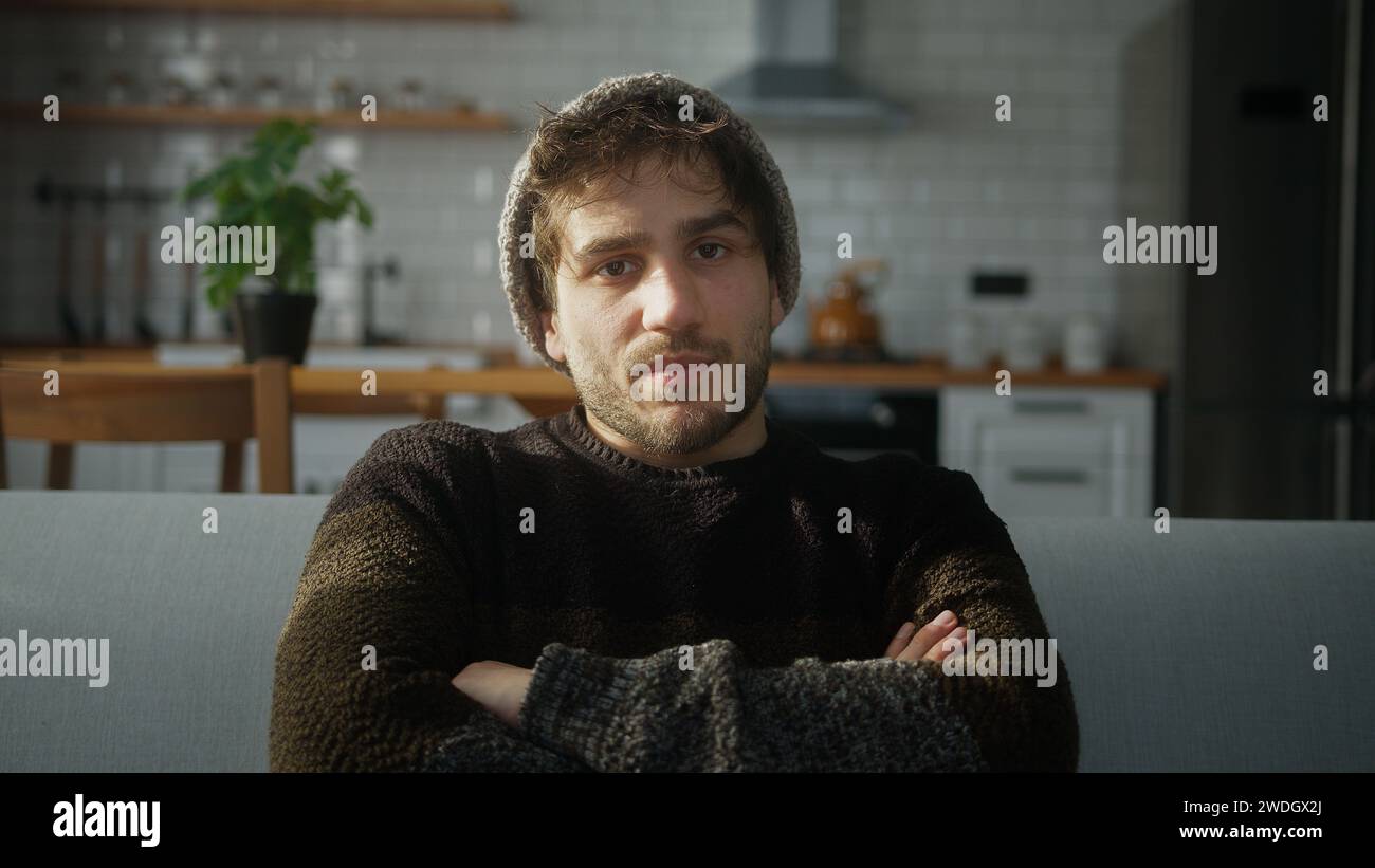 Bored young man with beanie sitting on sofa at home with kitchen background, having a conference call, listening to the speaker on the video call Stock Photo