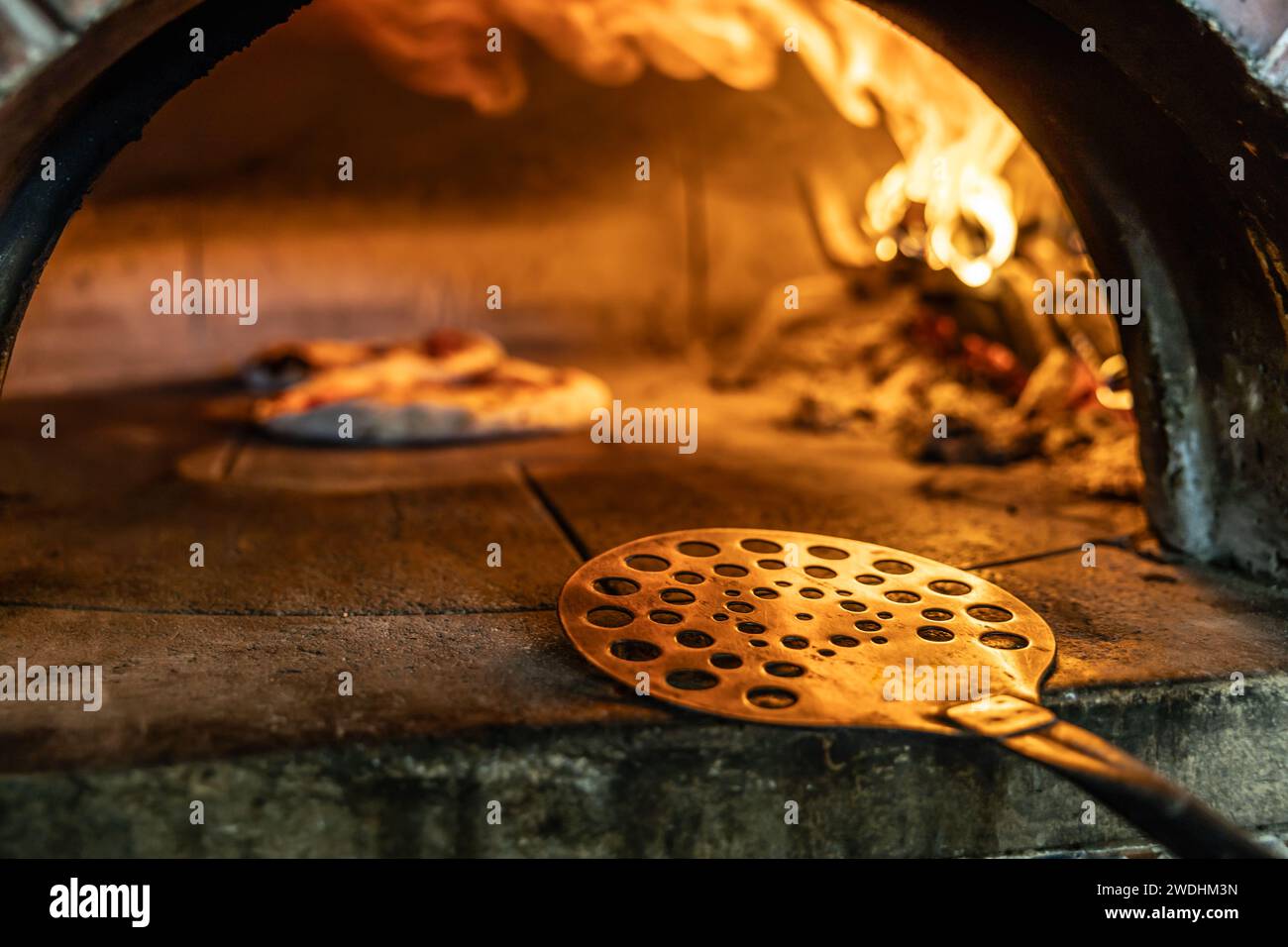 Traditional oven for baking pizza with burning wood and shovel. A stainless steel pizza shovel placed on the edge of a brick oven. Stock Photo