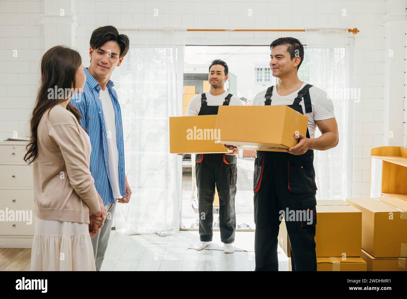 Professional movers aid couple indoors unloading boxes from truck into the home. Service guarantees Stock Photo