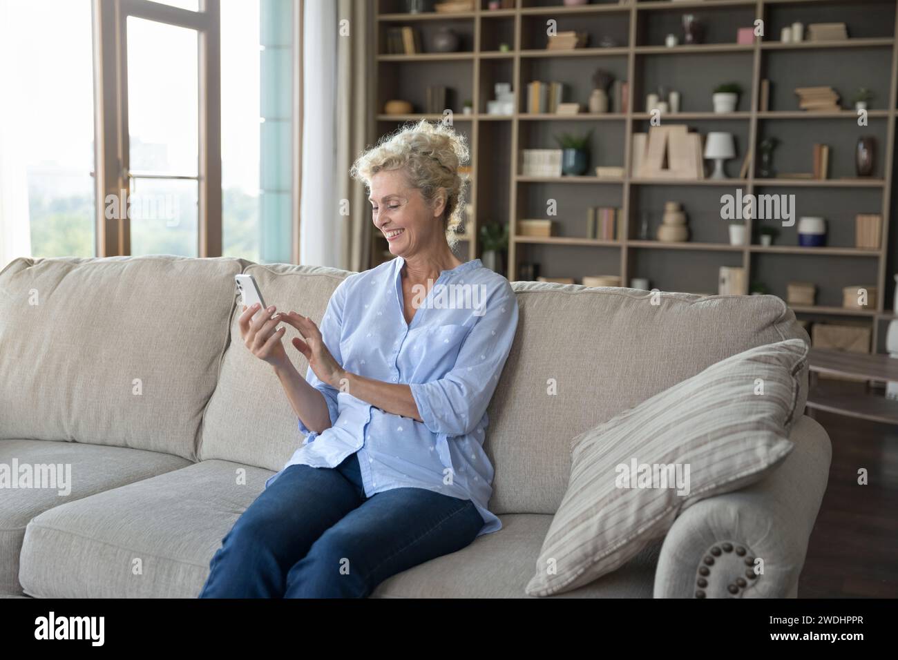 Mature woman enjoys chatting on-line sit on sofa with smartphone Stock Photo