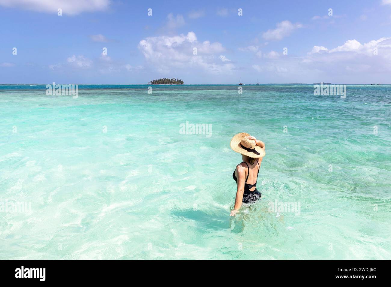Woman tourist with a straw hat walking in swimsuit in a turquoise shalllow water with a tropical island at the back, San blas, Caribbean sea, Panama Stock Photo