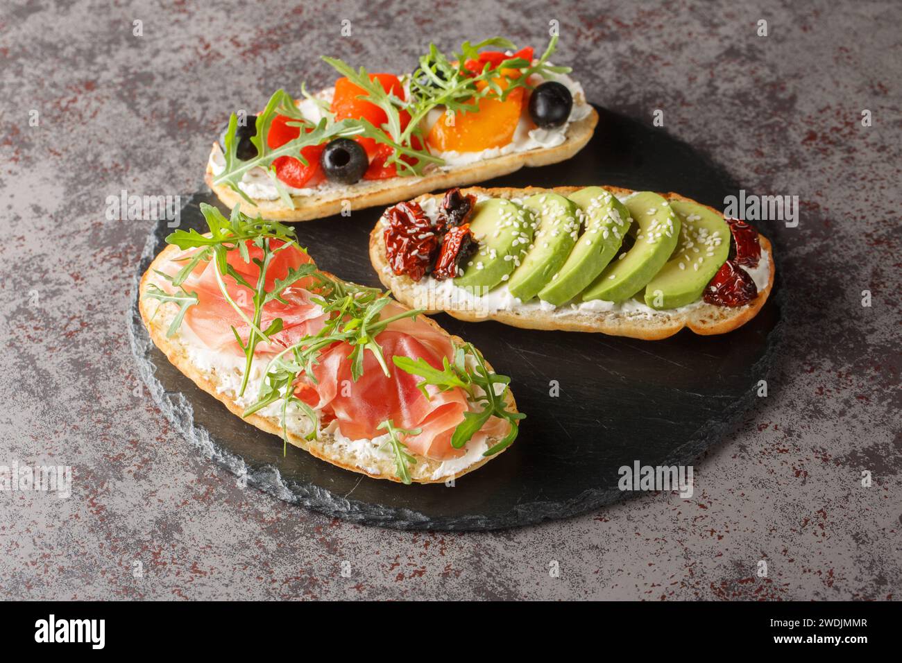Delicious bruschetta with arugula, ham, vegetables, cream cheese close-up on a slate board on the table. Horizontal Stock Photo