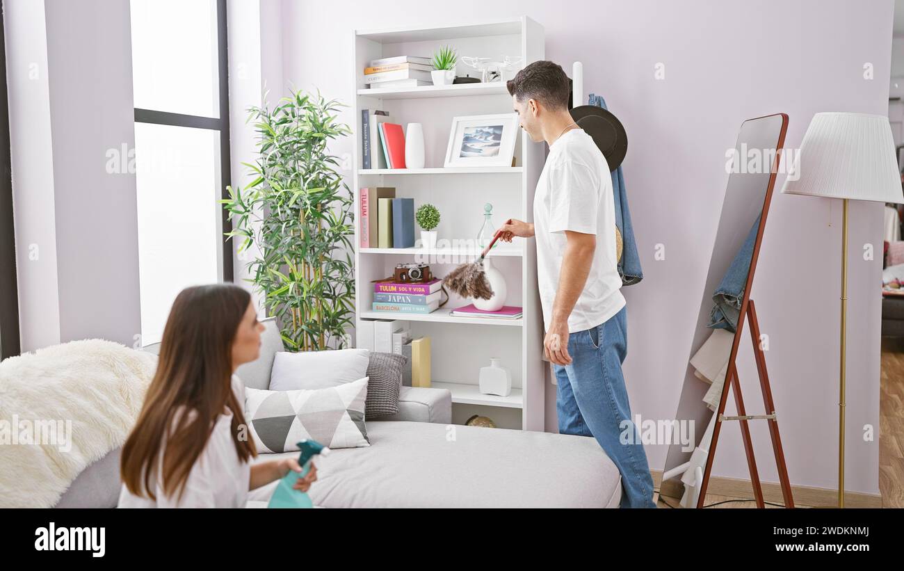 A woman sprays a sofa while a man dusts books in a well-lit modern living room, showcasing domestic life and teamwork. Stock Photo