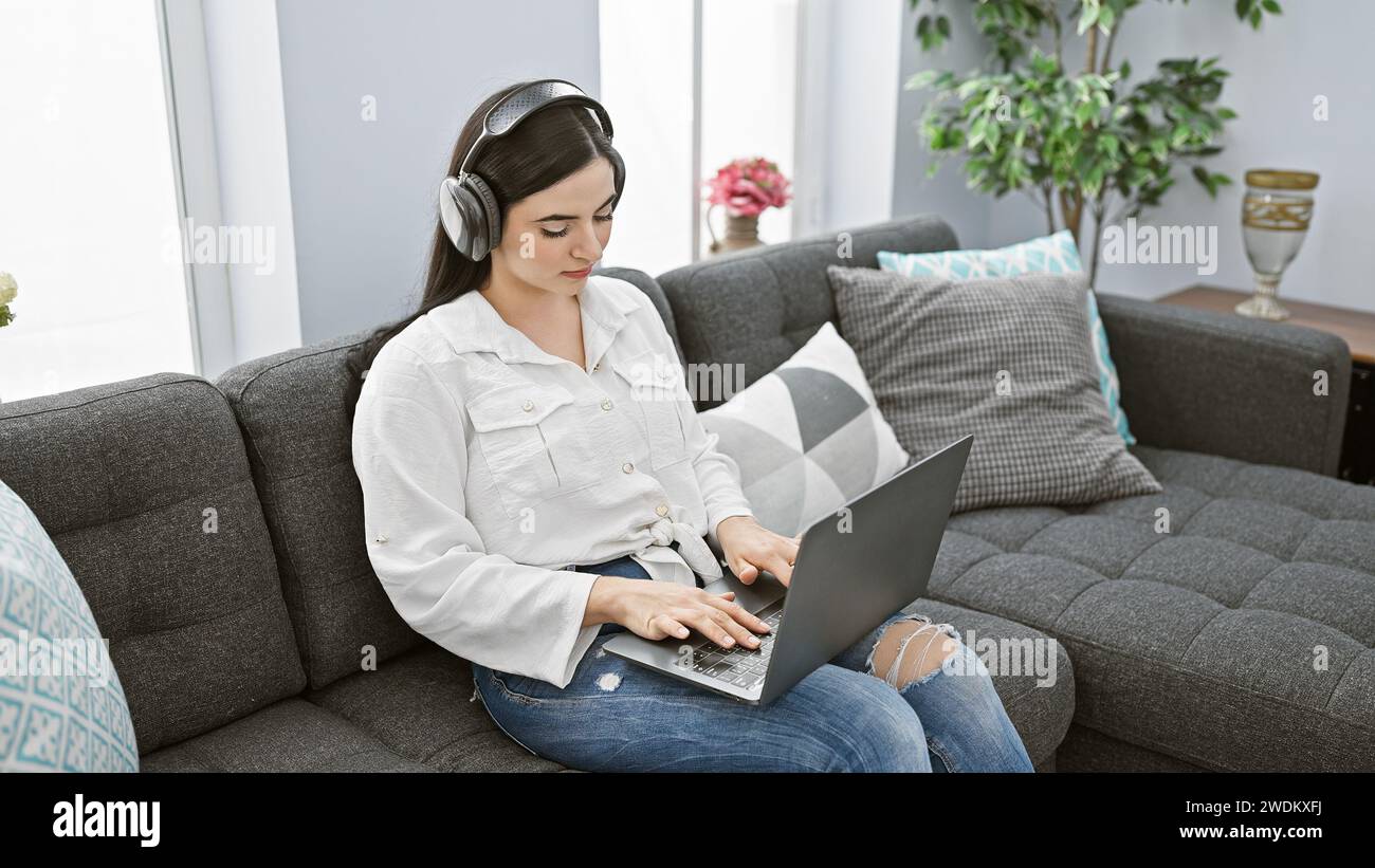 A focused hispanic woman wearing headphones uses a laptop while seated ...