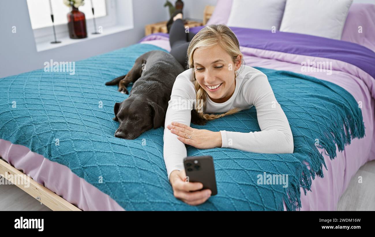 A young woman lies beside her labrador on a bed, taking a selfie in a ...