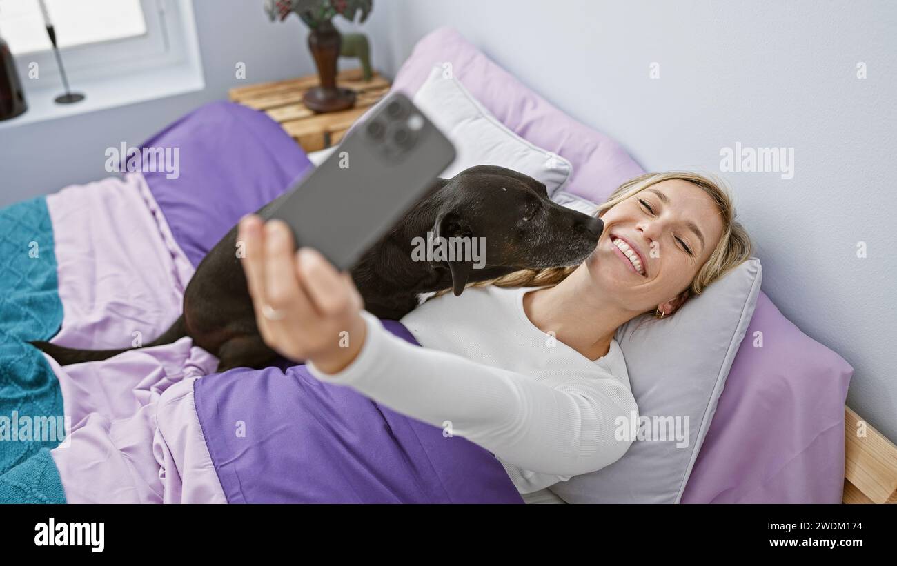 Blonde woman takes a selfie with her black labrador in a cozy bedroom ...