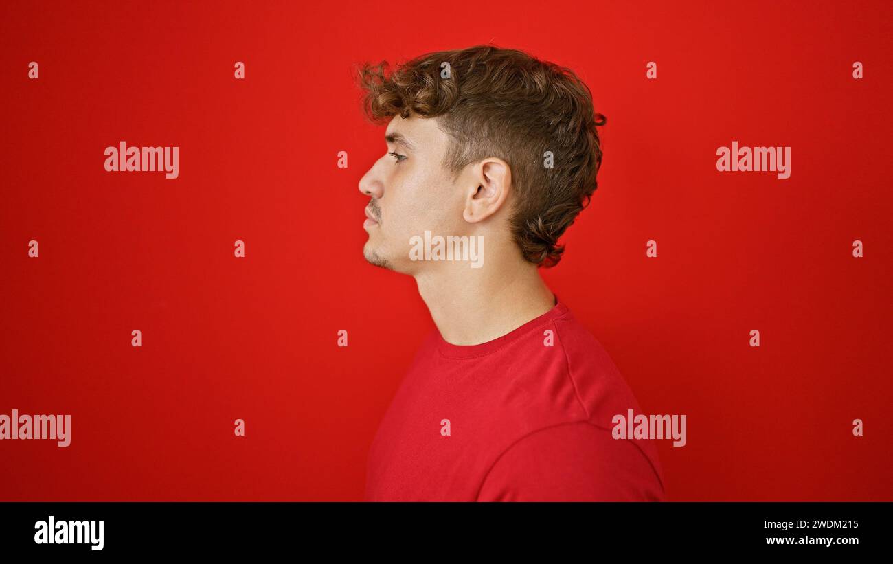 Cool, young hispanic man giving a serious side glance standing in the sunny light, portraying a relaxed lifestyle over an isolated red wall background Stock Photo