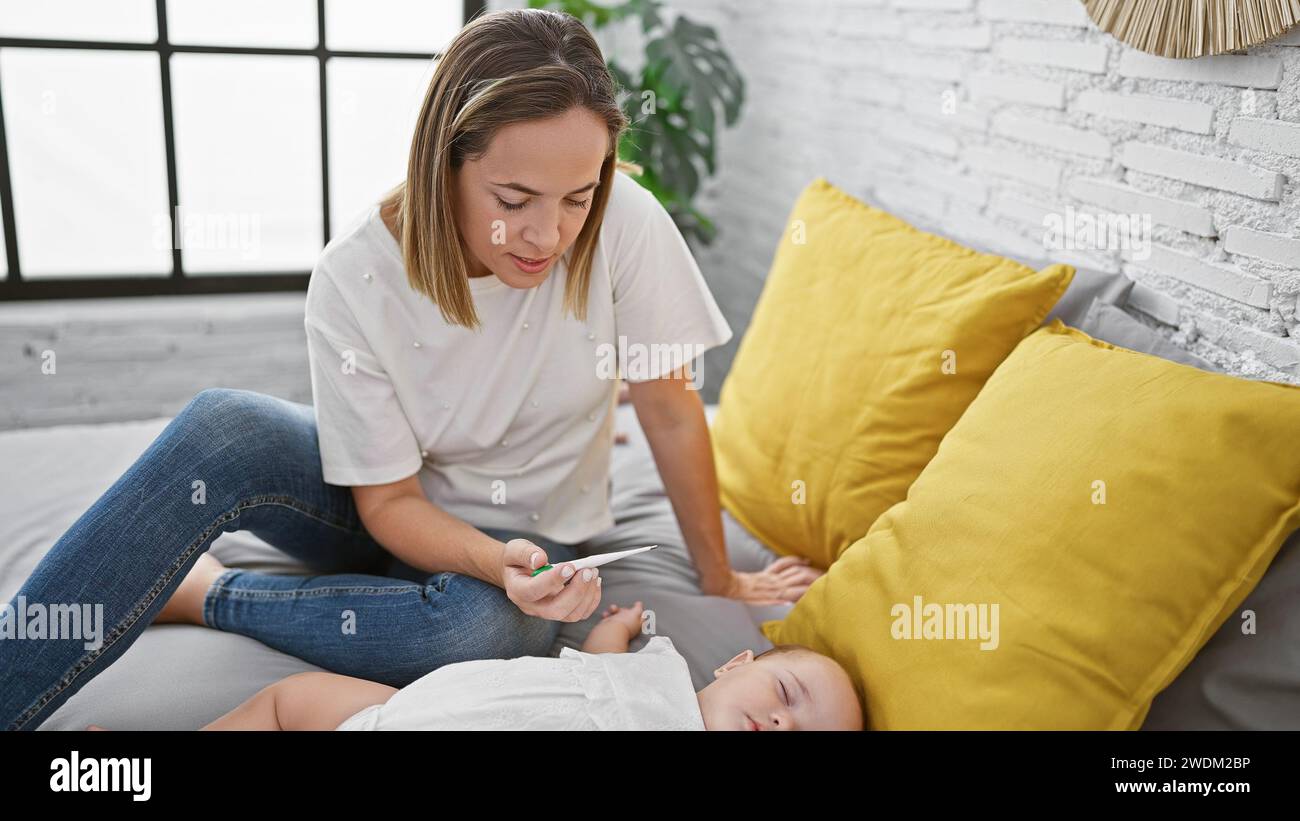 Loving mother measuring her sleepy daughter's temperature with a thermometer while she rests on the bed in their cozy bedroom Stock Photo