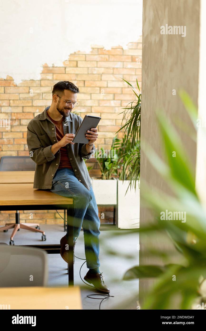 Cheerful man stands confidently holding a digital tablet in a contemporary office space with an exposed brick wall, symbolizing a blend of modern tech Stock Photo