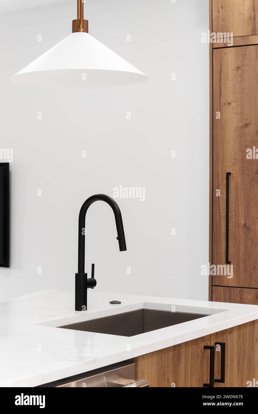A kitchen faucet detail with wood cabinets, a black faucet, and gold light fixture hanging above the  marble countertops. Stock Photo