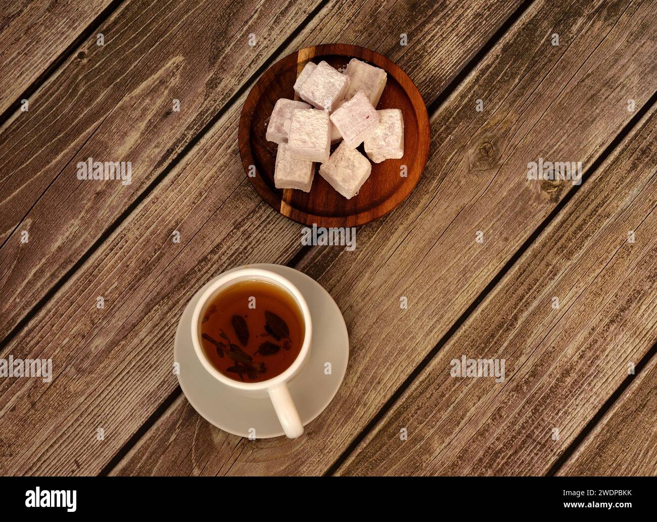 A white porcelain cup of hot tea on a saucer and a round wooden plate with a bunch of oriental sweets in powdered sugar on a wooden table. Top view, f Stock Photo