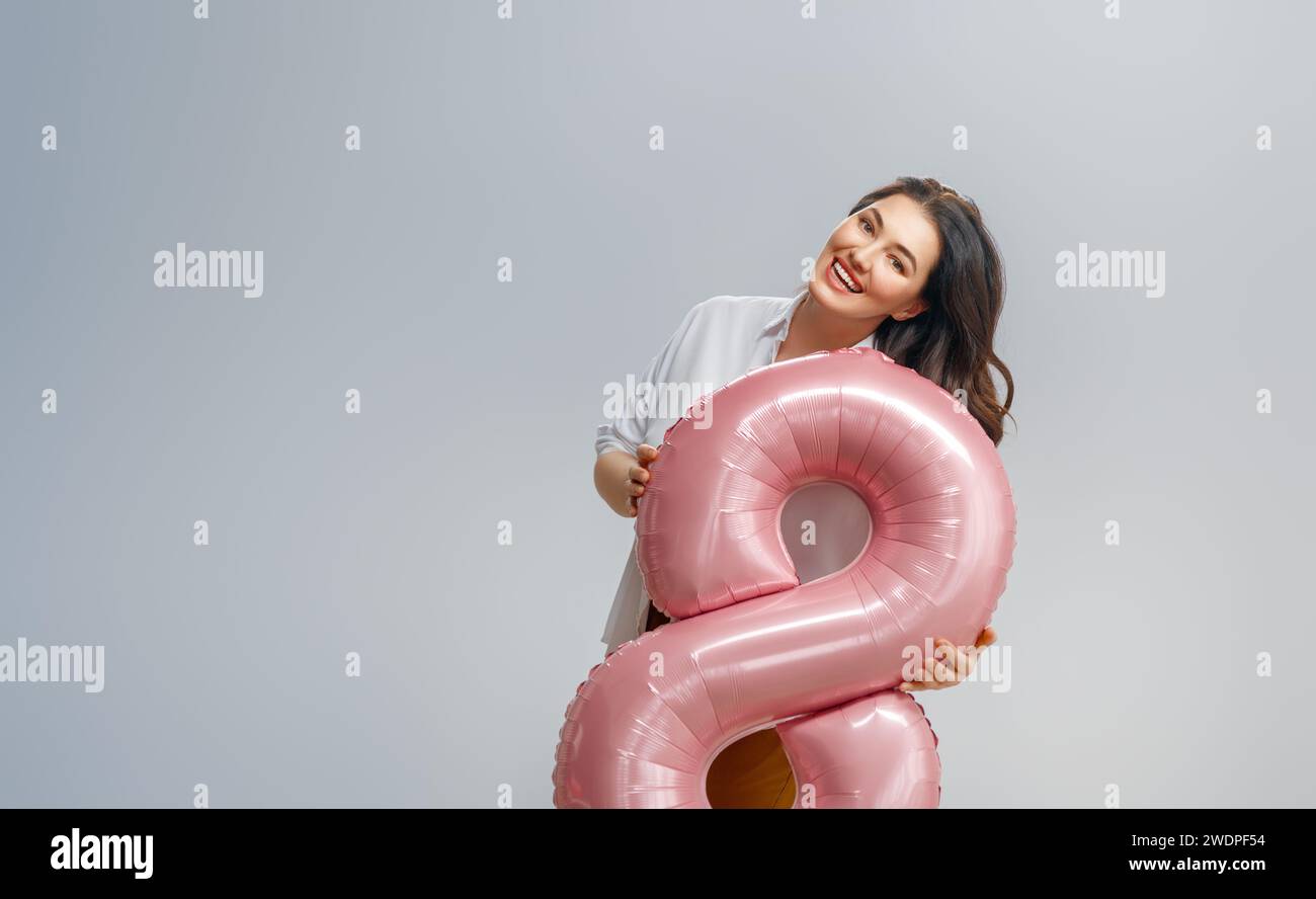 Beautiful young woman with a balloon in the shape of an eight on gray wall background. Stock Photo