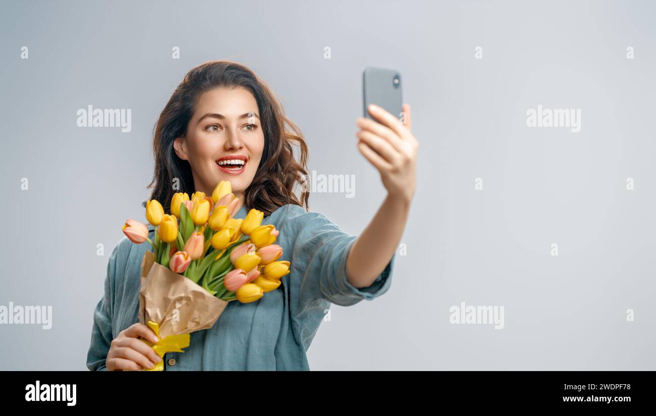 Beautiful young woman with yellow flowers in hands on grey wall background taking selfie. Stock Photo