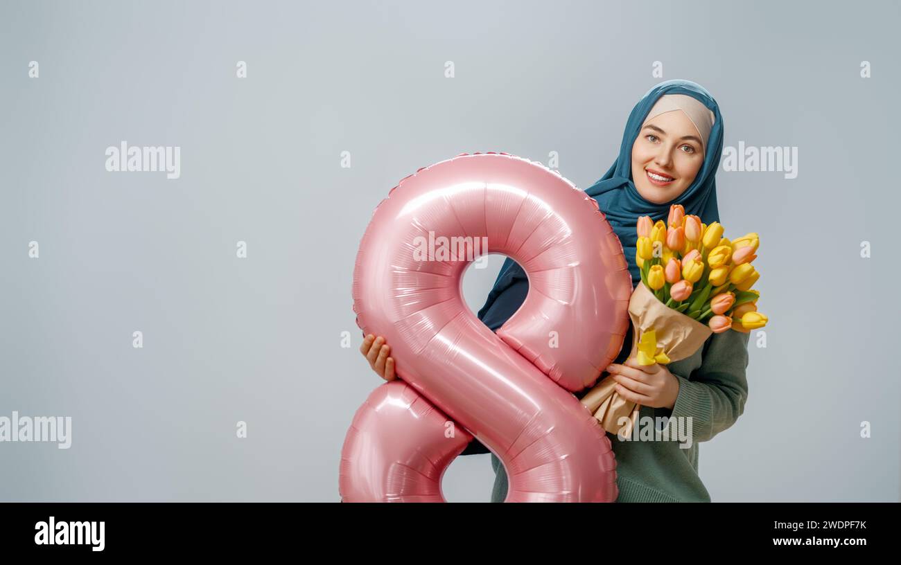 Beautiful young woman with yellow flowers and a balloon in the shape of an eight on gray wall background. Stock Photo