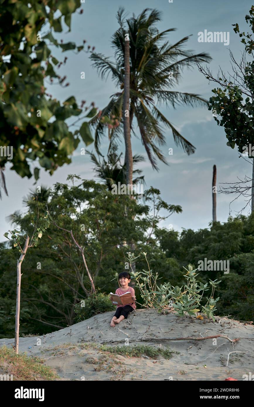A cute little girl sits under a tree reading a book in the tropical ...