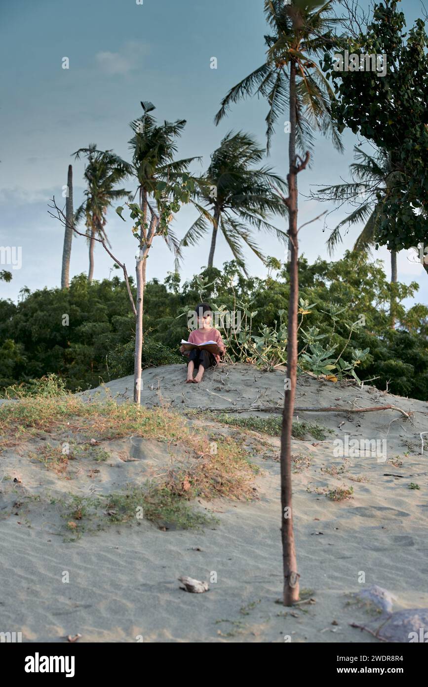 A cute little girl sits under a tree reading a book in the tropical ...