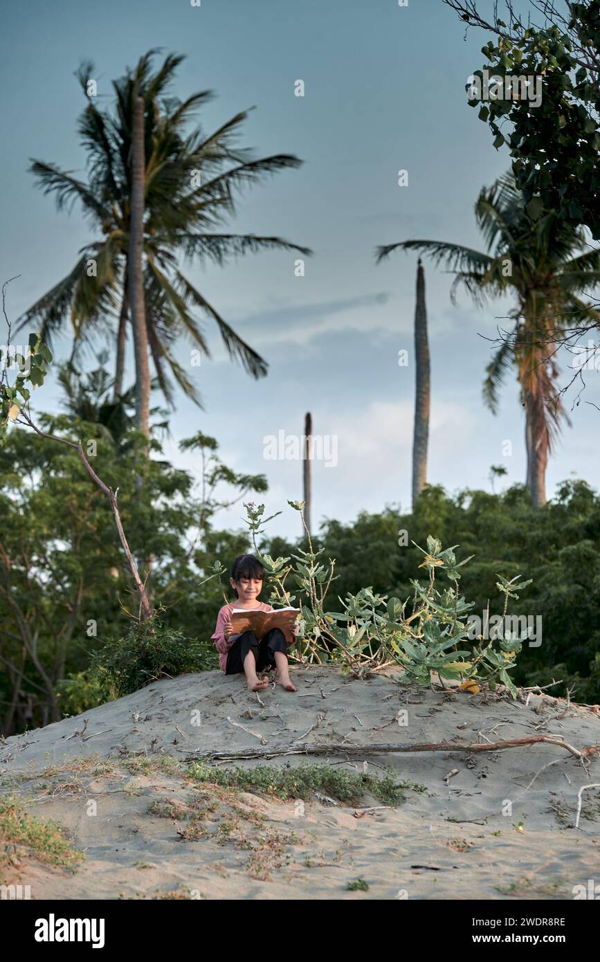 A cute little girl sits under a tree reading a book in the tropical ...