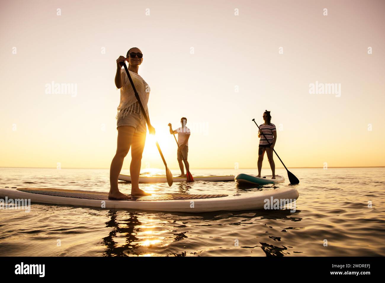 Three active young tourists are walking on standup paddle sup boards at sunset lake Stock Photo