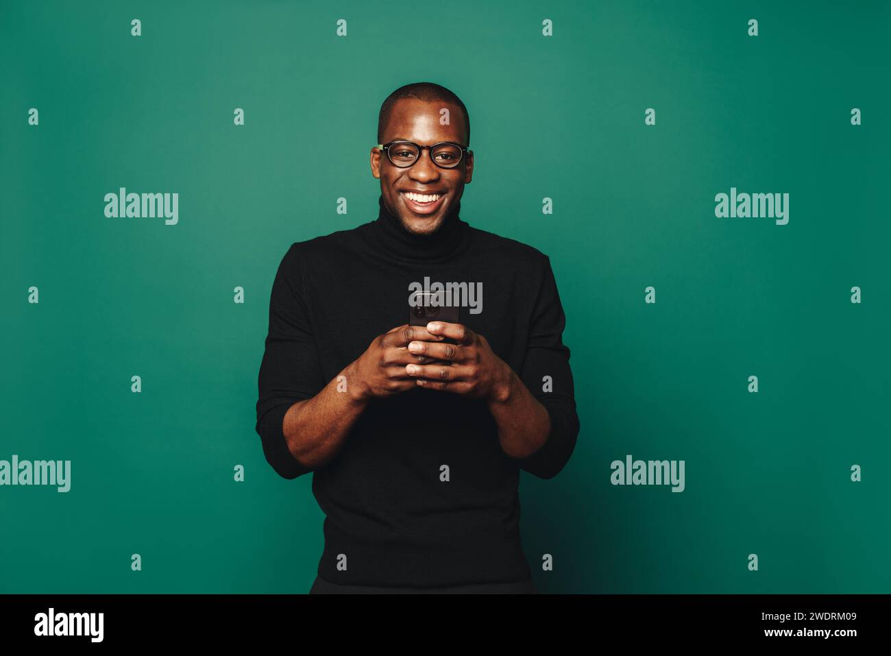 Portrait of a smiling young man in casual clothing standing against a green background, holding a smartphone. He stays connected and social with this Stock Photo