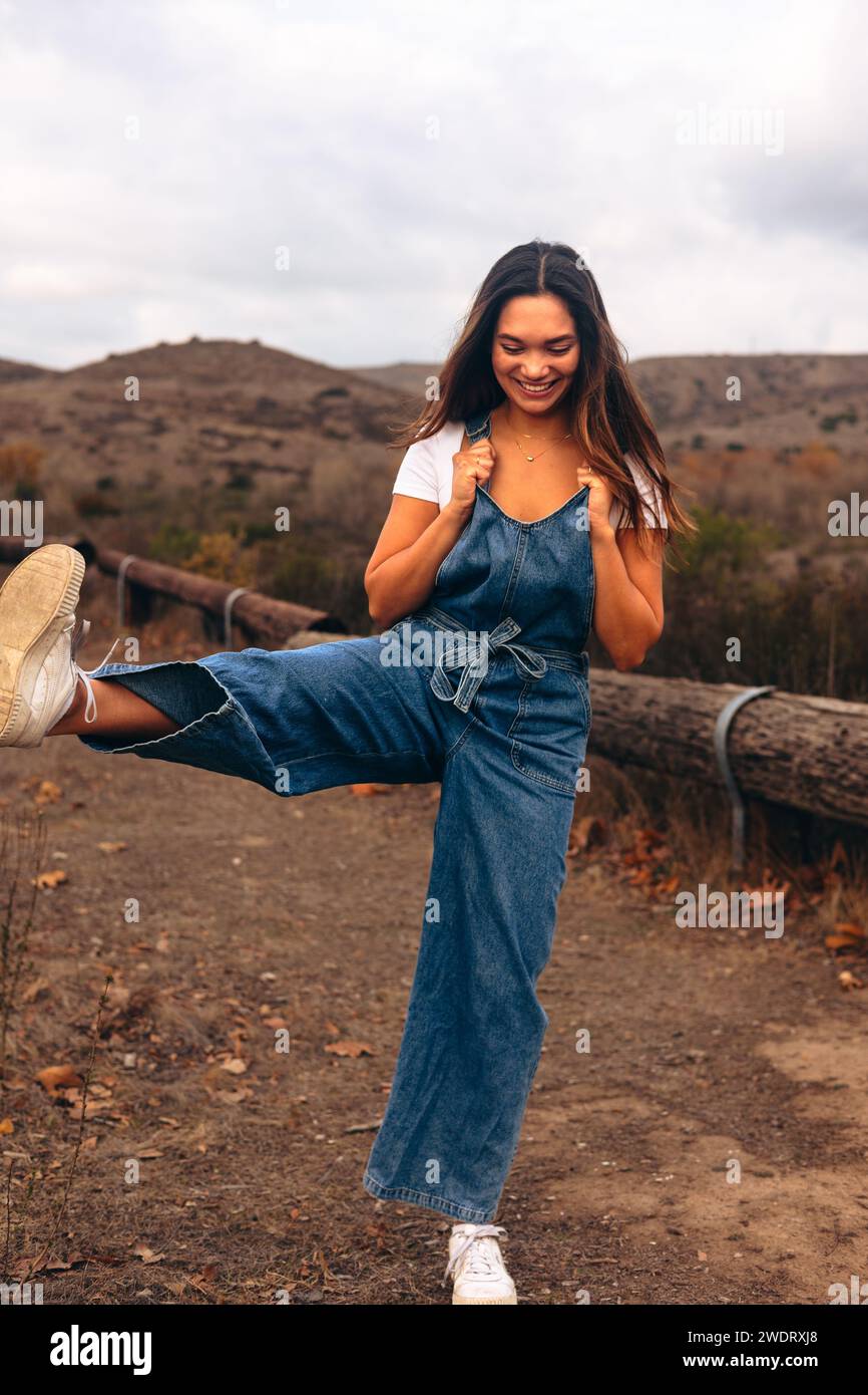 Young woman wearing overalls kicking air on a hiking trail Stock Photo