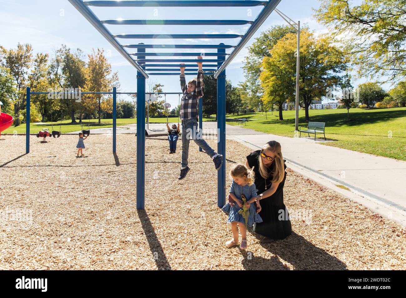 Brother does monkey bars while mom helps sister with dinosaur toy Stock Photo