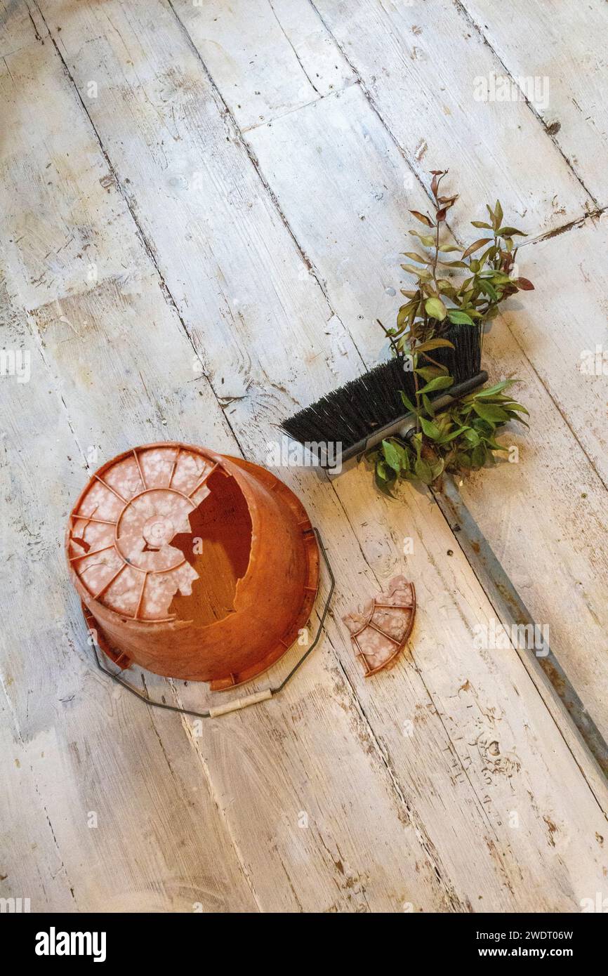 An upturned orange plastic bucket with an enormous hole in its bottom and a broom with greenery growing round it on a white washed floor Stock Photo