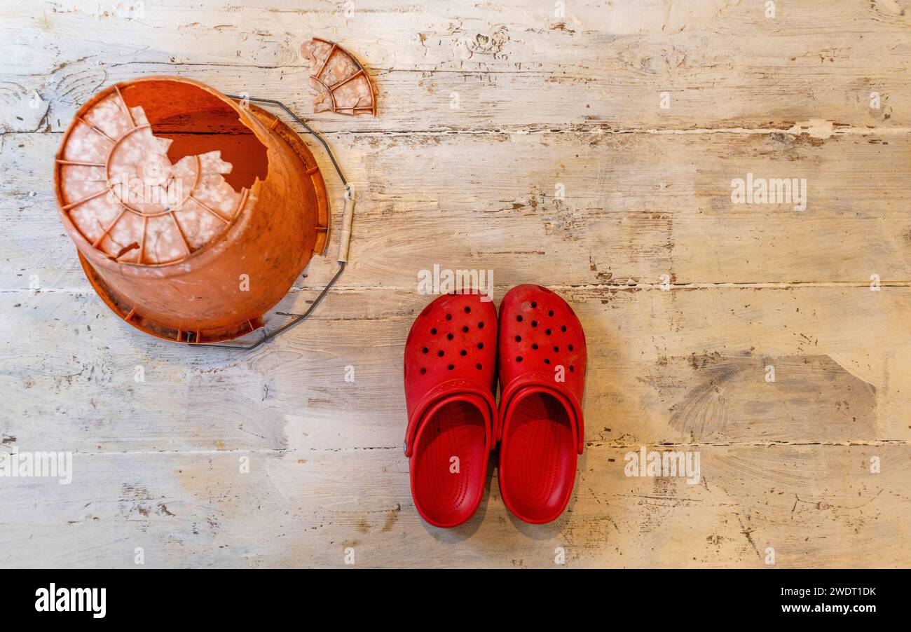 On whitewashed floorboards an inverted orange plastic bucket with an enormous hole in the bottom and a pair of bright red Crocs Stock Photo