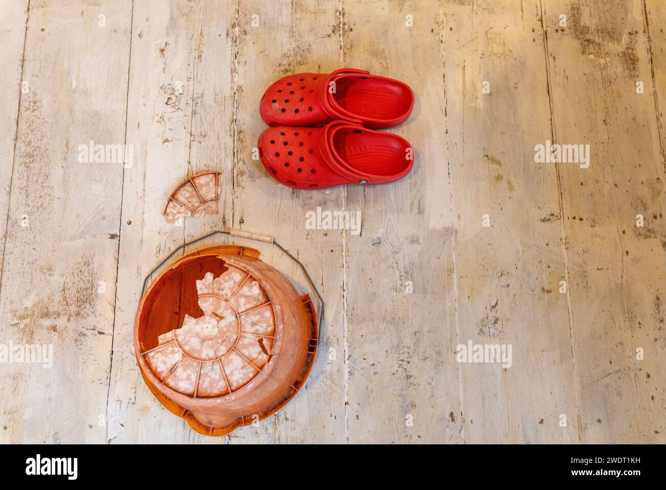 On whitewashed floorboards an inverted orange plastic bucket with an enormous hole in the bottom and a pair of bright red Crocs Stock Photo