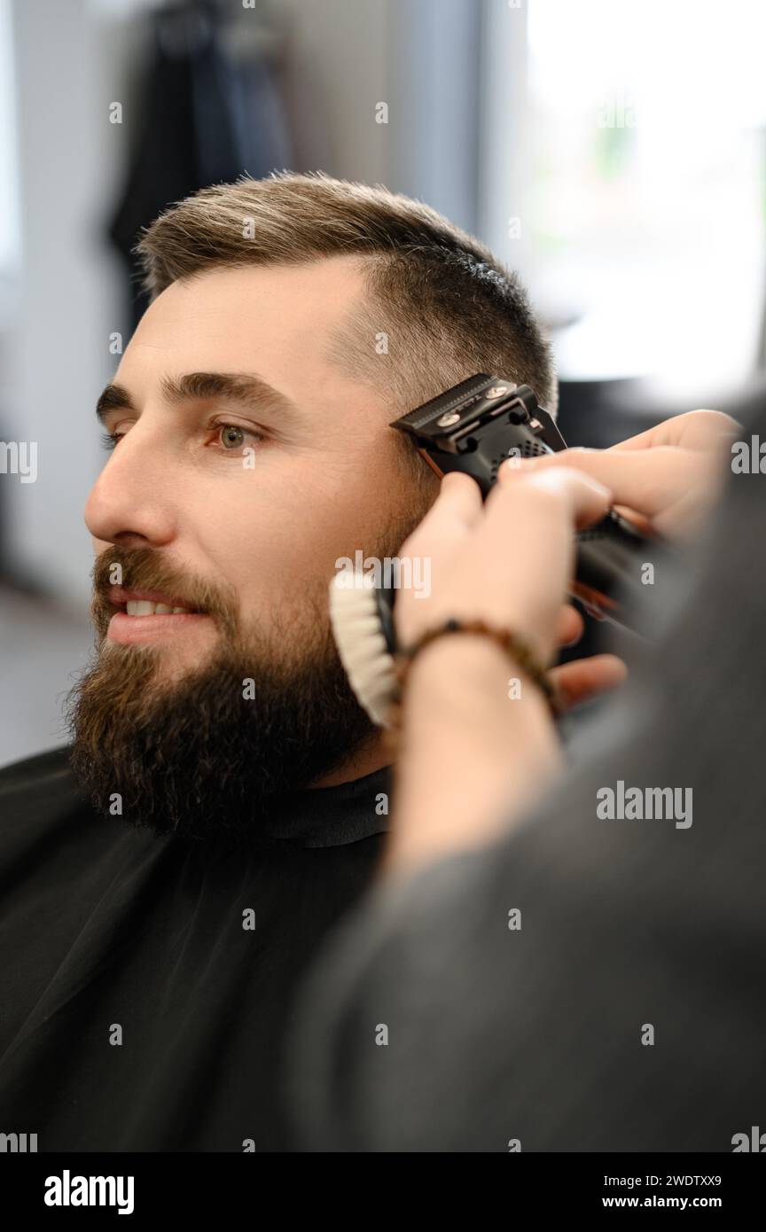 Barber shaves the contour of the oval line with a clipper on the clients head. A man with a beard gets a haircut in a barbershop chair. Stock Photo