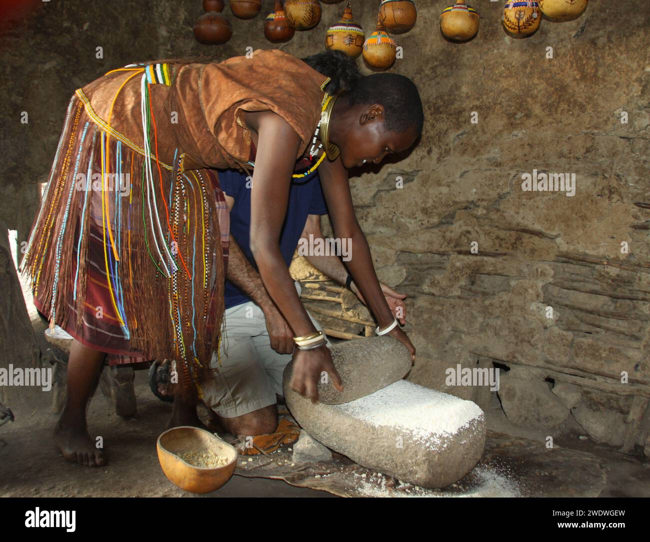 Datoga woman in traditional leather dress adorned with beads and brass bracelets milling grain to flour. Stock Photo