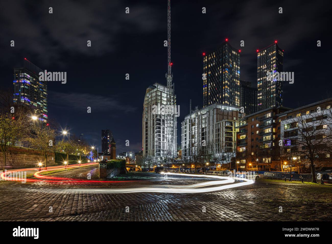 Image on a cobbled Castlefield car park at night in Manchester city centre, UK with car light trails, Beetham Tower, Castle Wharf & Deansgate Square Stock Photo