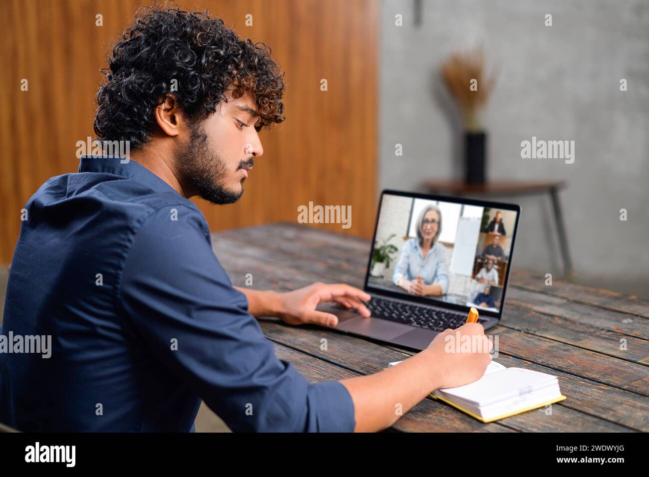 Focused young man with curly hair is participating in virtual meeting, taking notes as listens to a woman on his laptop screen. Modern remote work environment and the concept of digital communication Stock Photo